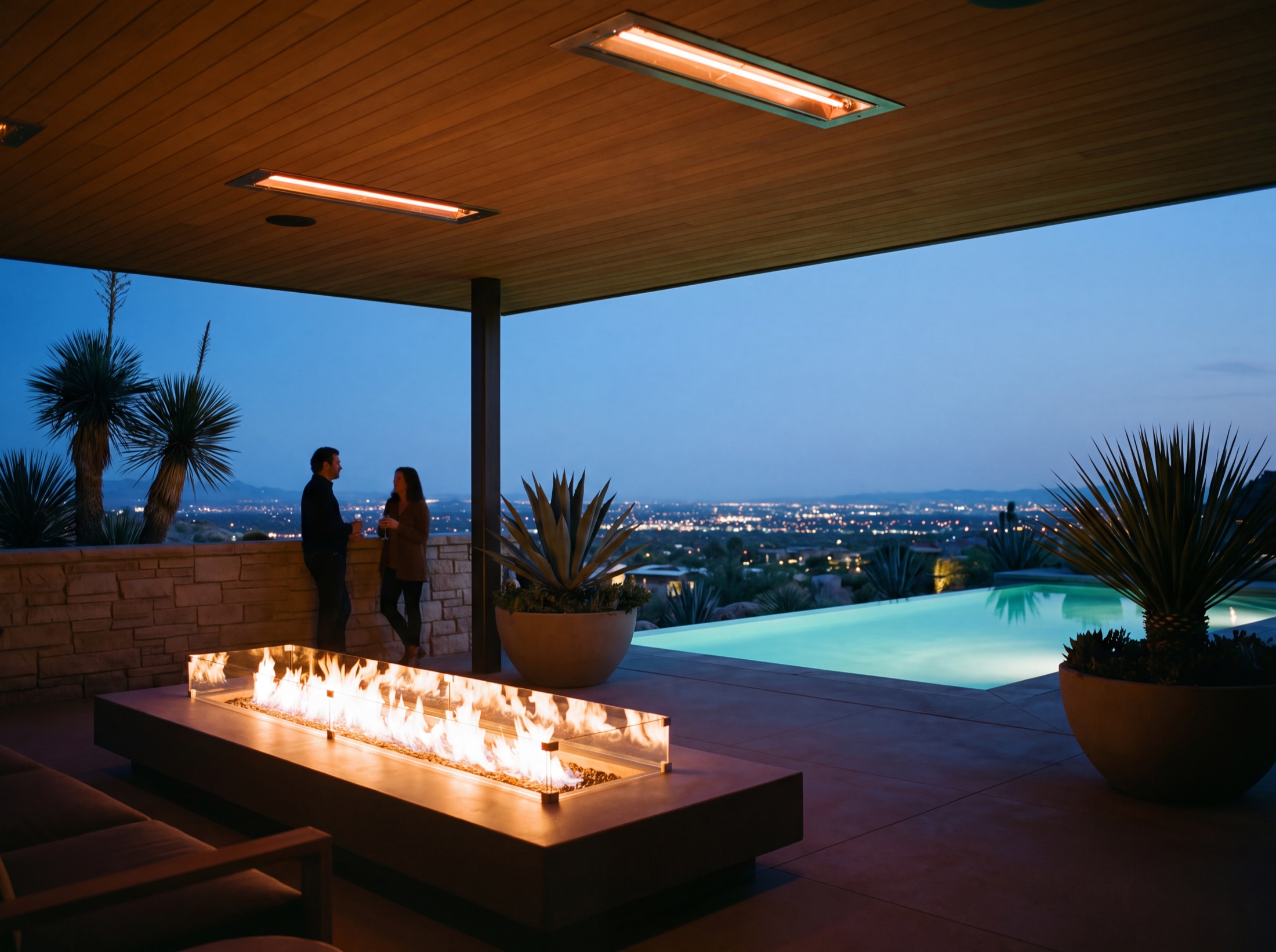 Covered outdoor living room at dusk, fire feature lit, the pool luminous beyond and the valley lights low in the distance