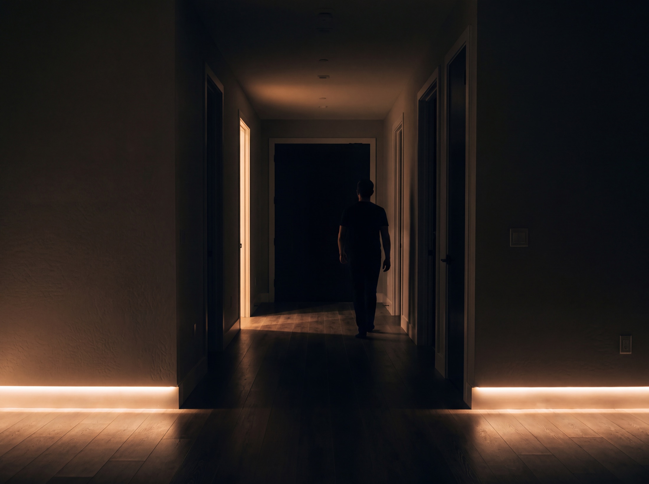 A hallway at night settling room by room, path lights at a low glow, the far rooms already dark