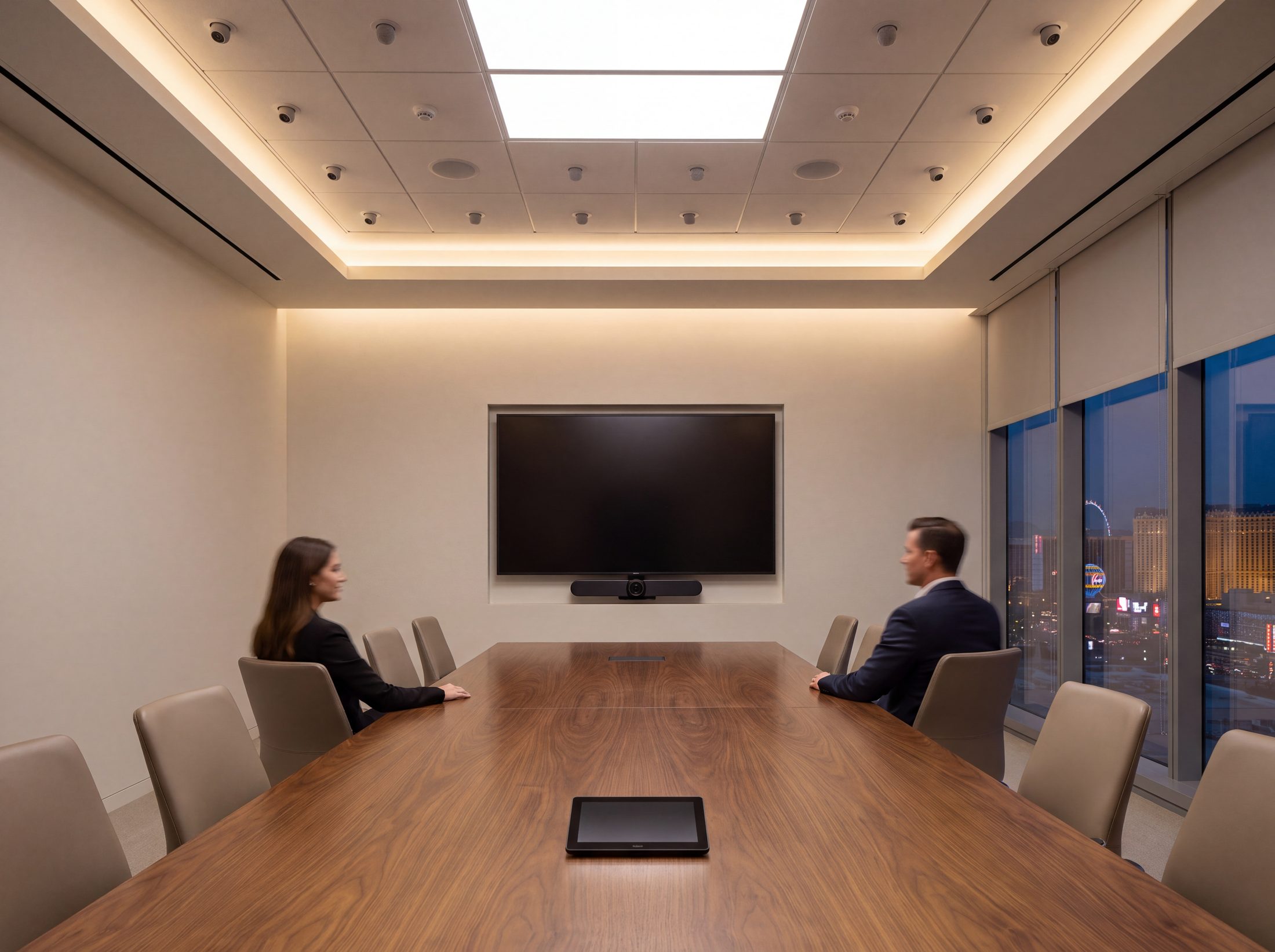 A hybrid meeting room seen down its center axis, the display and camera bar centered on the table line, two colleagues at the near end