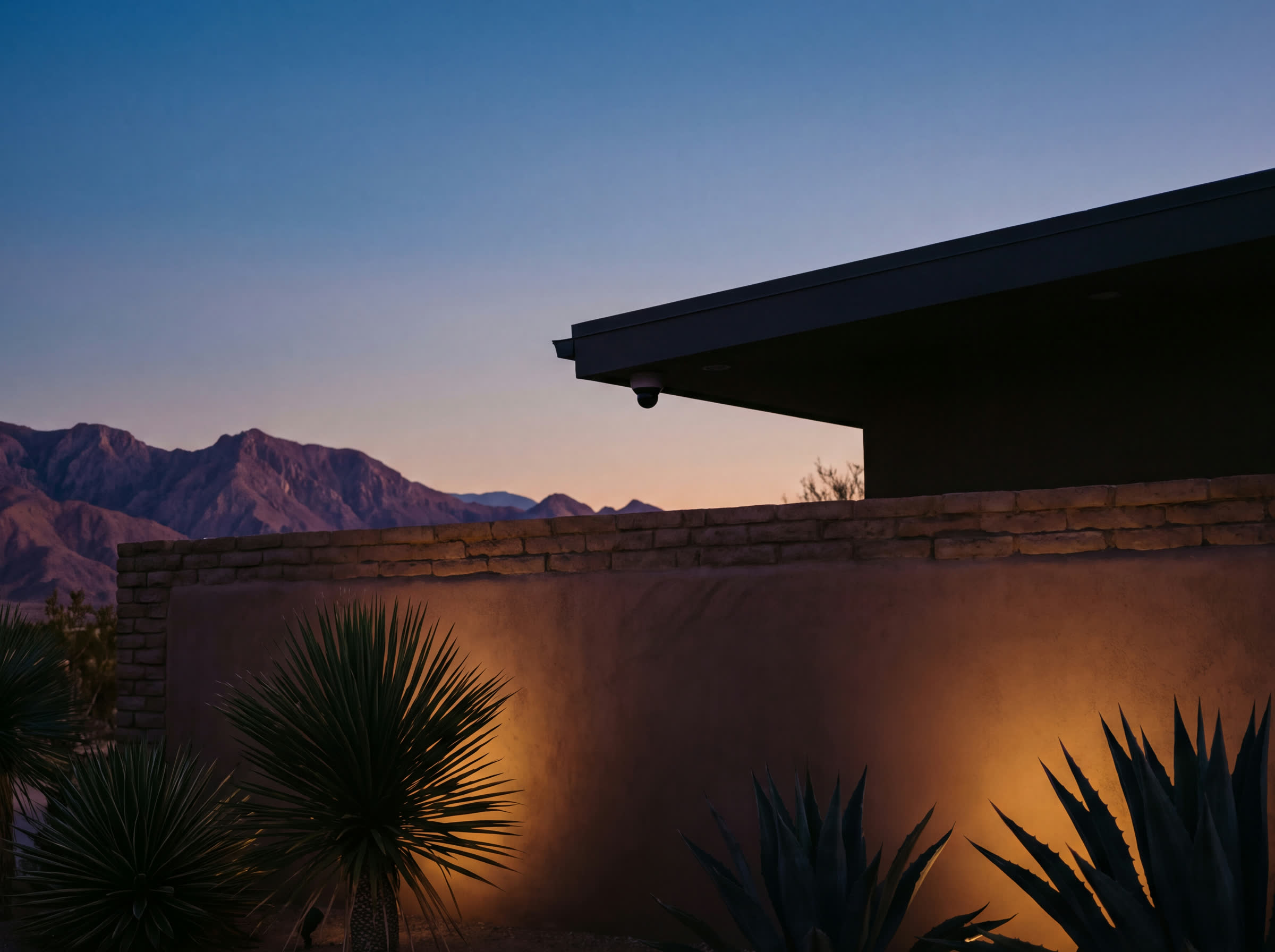 A dome camera tucked under the eave line at dusk, agaves uplit along the boundary wall, mountains behind