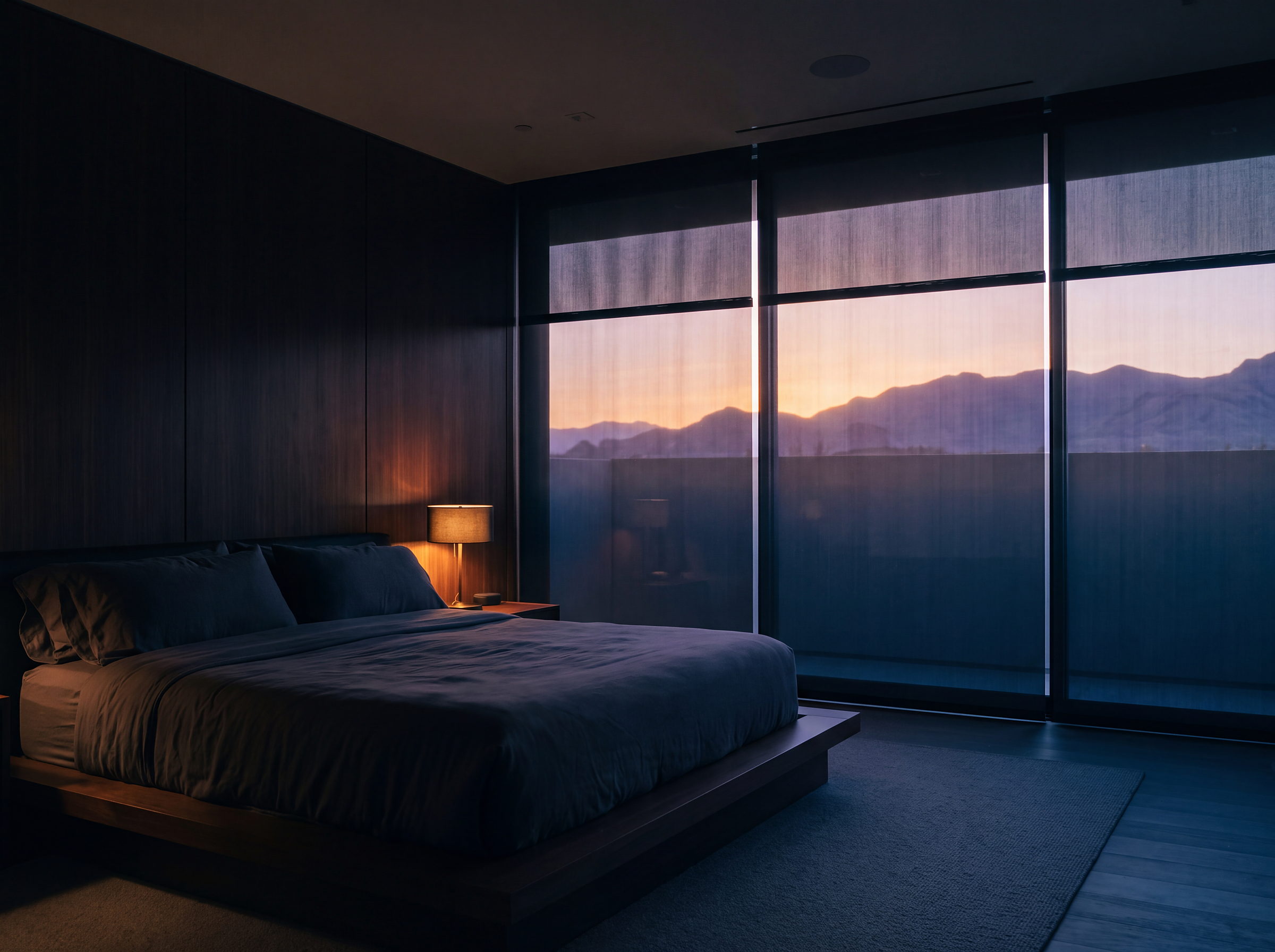 A bedroom at dawn, motorized shades part-lowered, first light on the desert ridges beyond