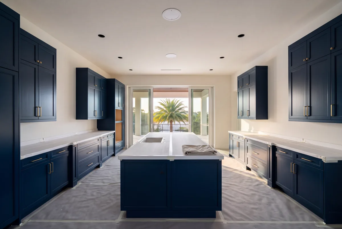 A new kitchen at trim-out with ink blue cabinets, countertops and floor covered in protective board and paper, and recessed light openings in the ceiling