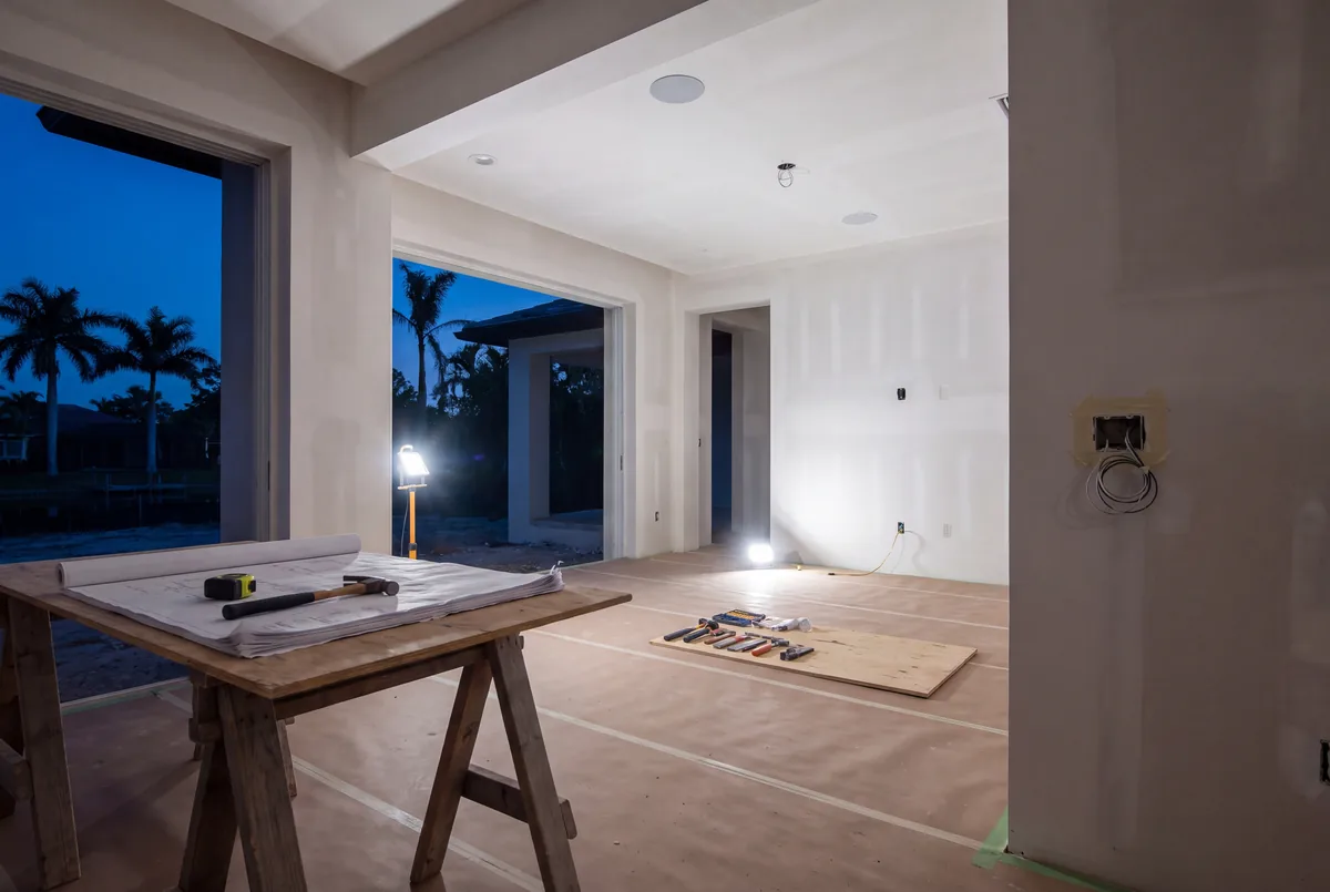 A living room before trim-out, the drywall hung, a wall box left open with its cable and the plans on a sawhorse table