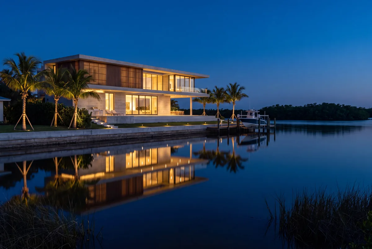 A new two story waterfront house at dusk across a still bay, lit inside, with step lights, path lights and uplit palms along the seawall