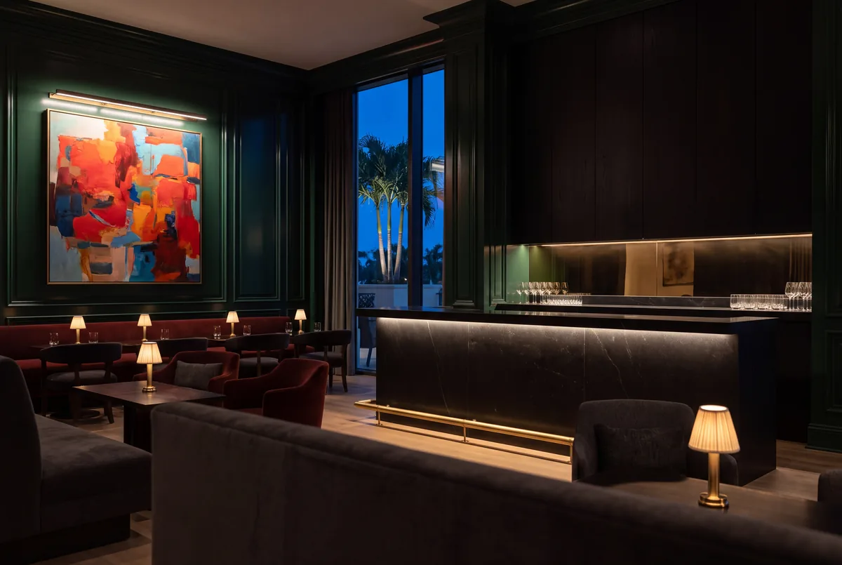 A club lounge at dusk with deep green paneling, a lit black stone bar with a brass rail, table lamps and a large abstract painting
