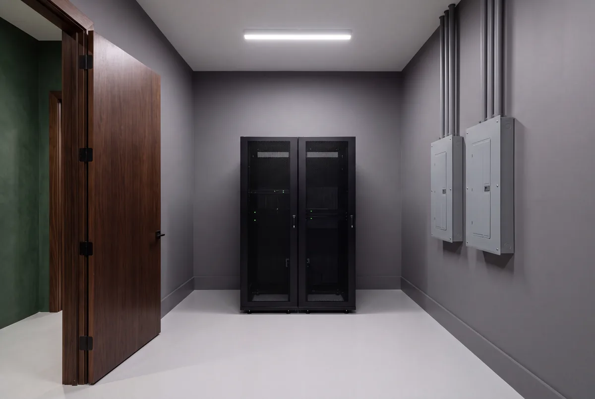 A clean graphite gray equipment room with two closed black equipment racks and two electrical panels on the side wall, seen through an open walnut door
