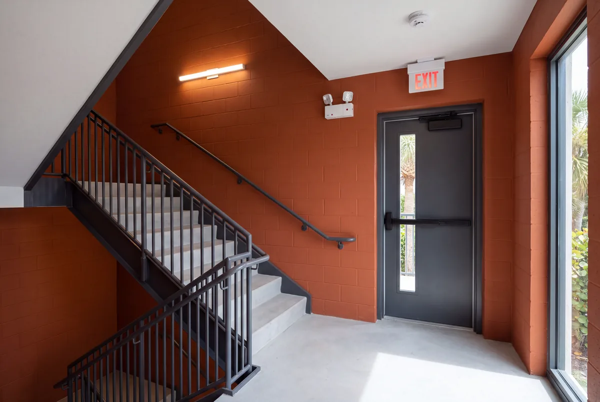 A stair landing with orange painted block walls, a red EXIT sign over the steel exit door, a twin head emergency light high on the wall beside it and a smoke detector on the ceiling