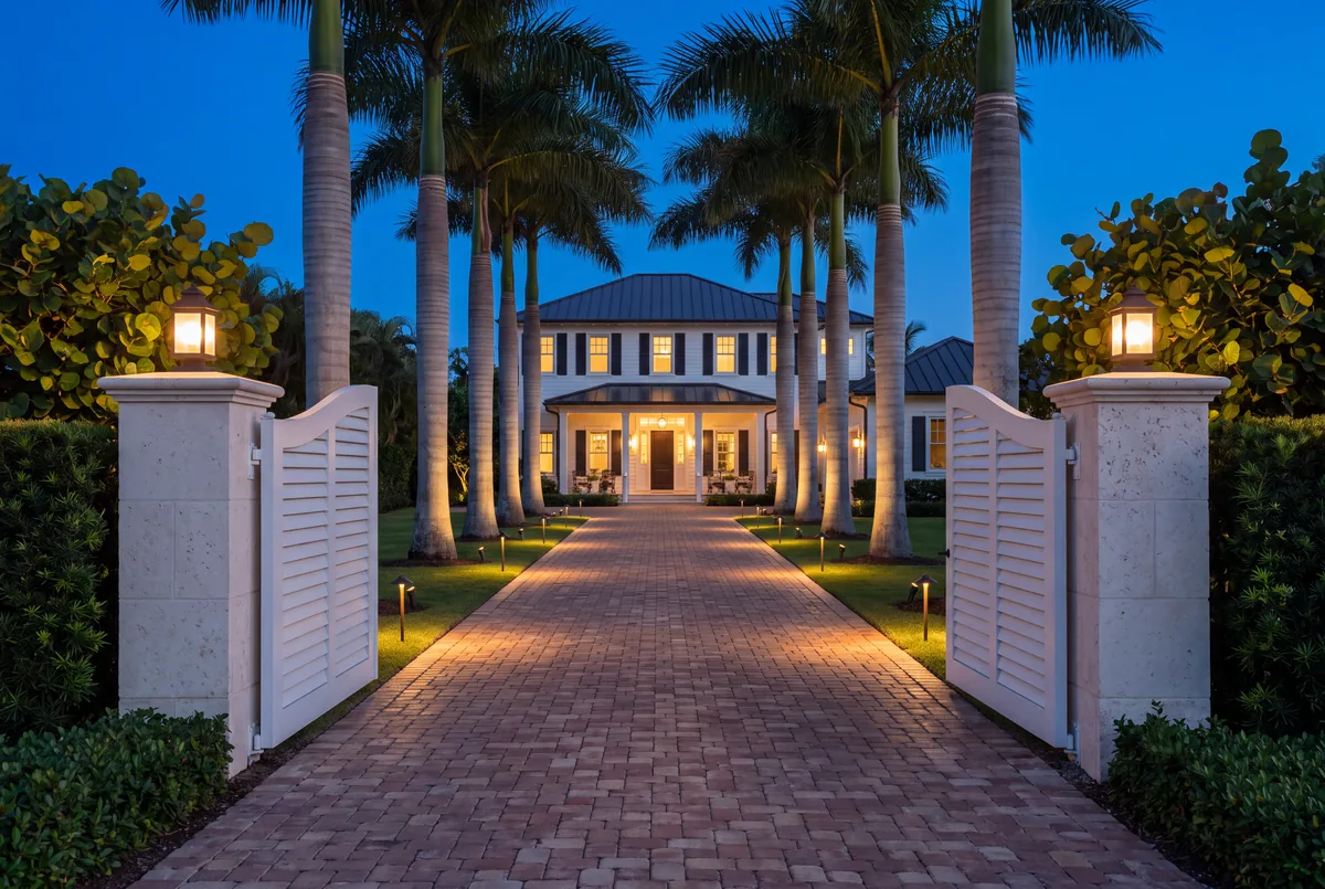 Open white louvered gates between coral stone piers at dusk, with path lights and uplit royal palms along a brick drive to a lit house