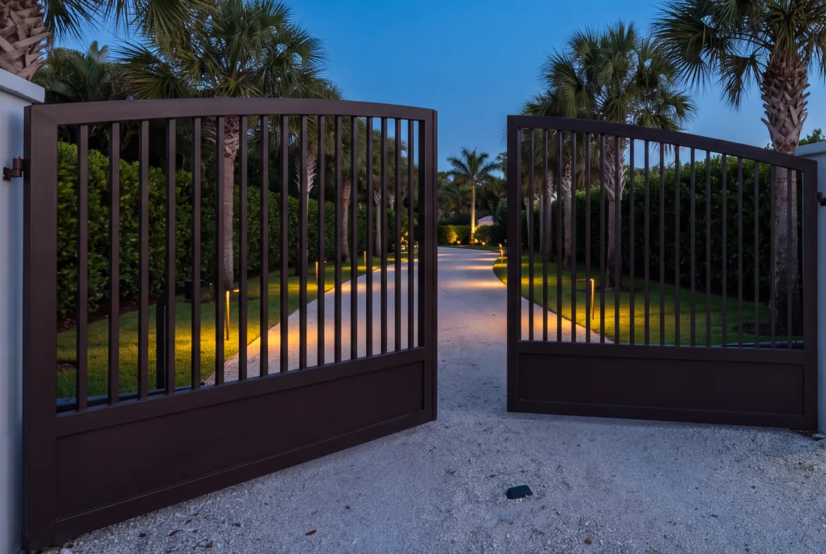 A pair of dark bronze driveway gates opening at dusk, path lights glowing along the drive between clipped hedges and sabal palms