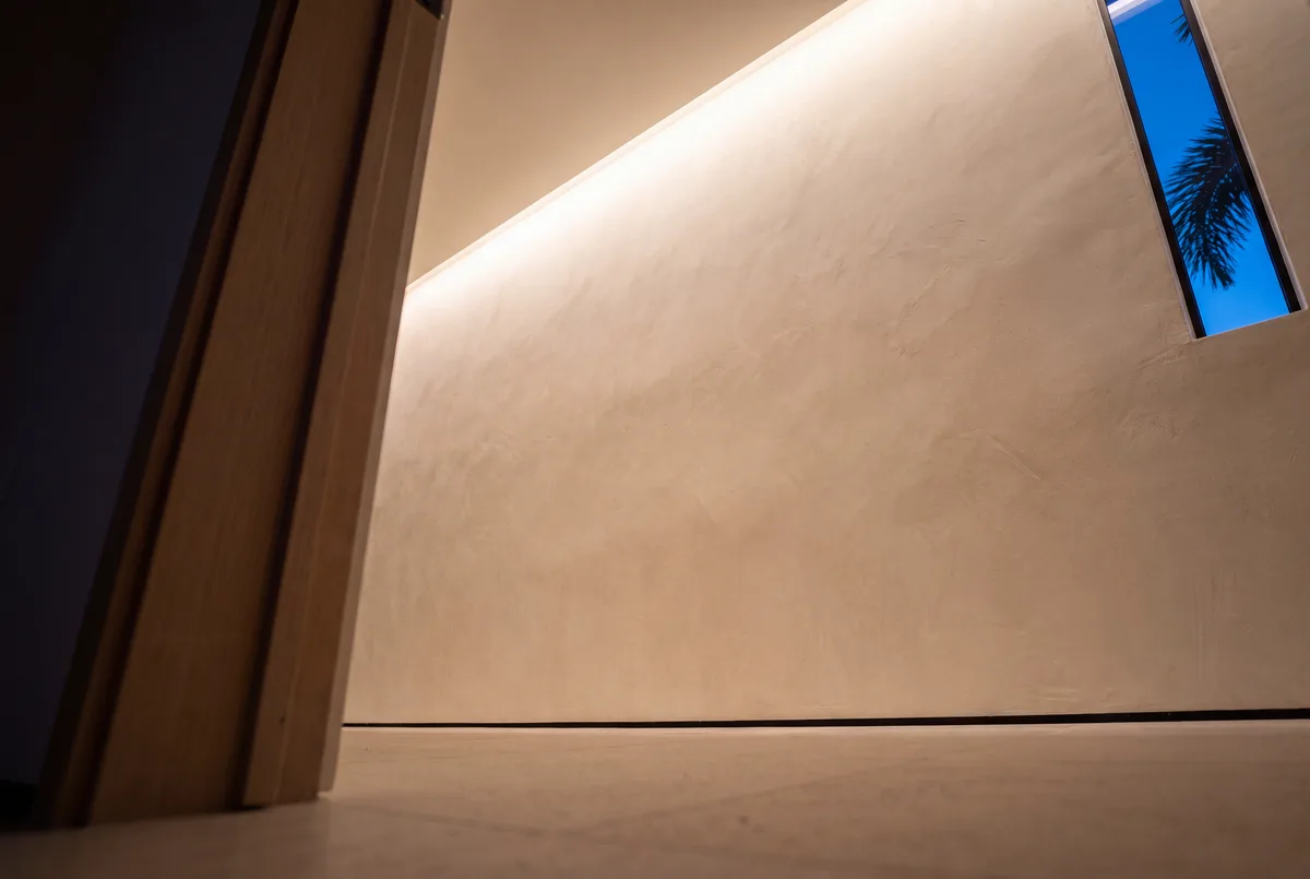 A ceiling slot washing a lime plaster wall with light that falls away evenly to the floor, the wall bare of switches