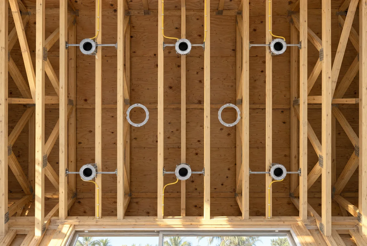 Looking up at ceiling framing before drywall, with six recessed light housings in two even rows of three and two round speaker brackets on the center line