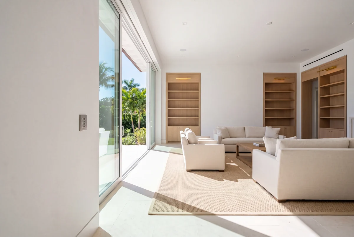 A bright coastal living room in white, oak and linen, sliding glass open to a garden, with a flush scene keypad beside the opening