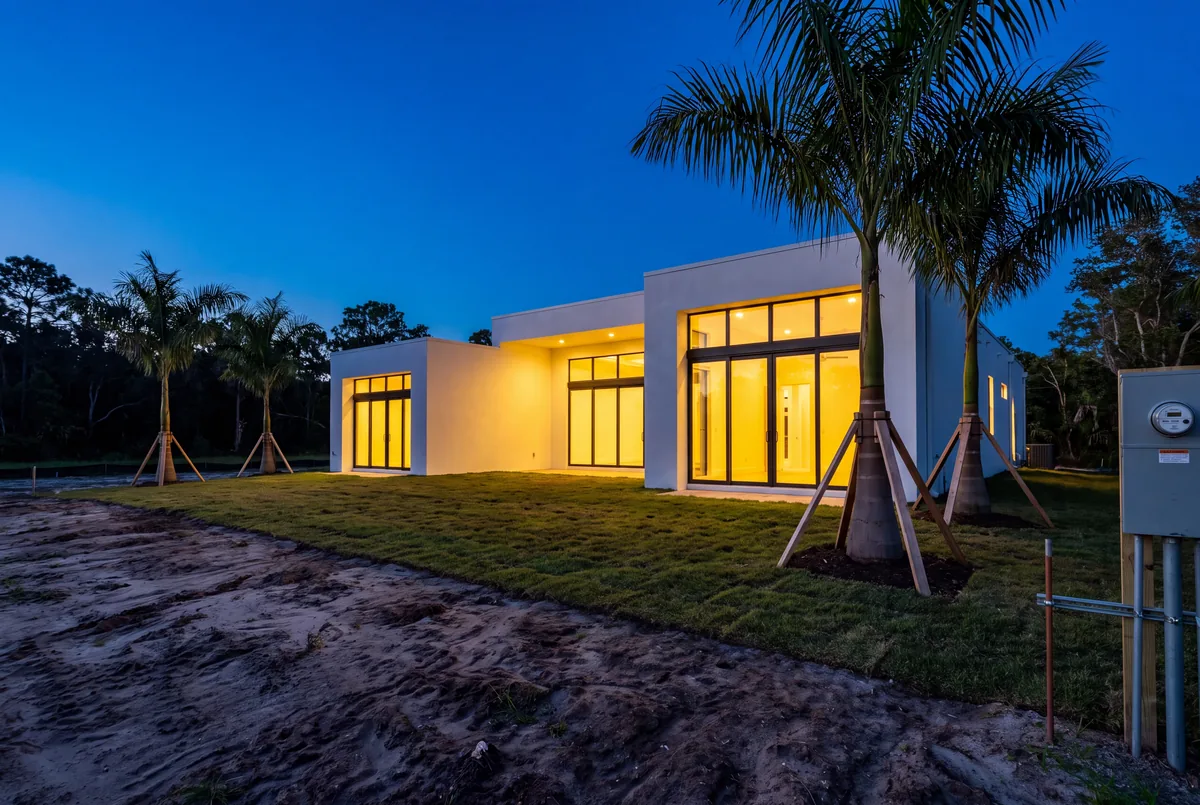 A modern house nearing completion at dusk, lit inside for the first time, with fresh sod and young palms on the site